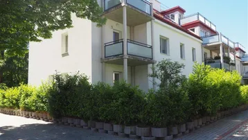 Weißes Mehrfamilienhaus mit Balkonen und gepflegter Hecke im Außenbereich unter blauem Himmel.