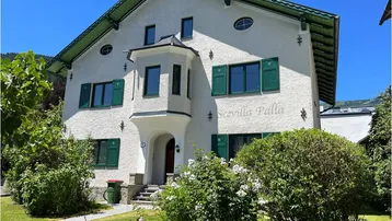 Charmante Seevilla mit weißer Fassade, grünen Fensterläden und gepflegtem Vorgarten unter blauem Himmel.