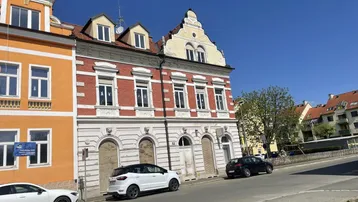 Historische Gebäude mit roten Ziegeln und orangefarbenen Fassaden entlang einer Straße mit geparkten Autos unter klarem Himmel.