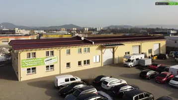 Luftaufnahme eines Gewerbegebäudes mit großem Parkplatz und Bergen im Hintergrund, inklusive Vermietungsschild.