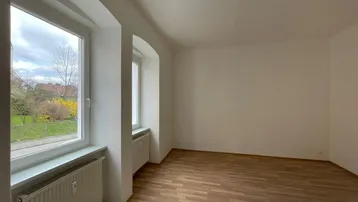 Helles Zimmer mit Laminatboden und Blick ins Grüne durch große Fenster.