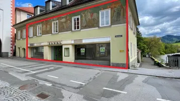 Historisches Gebäude mit kunstvollen Wandmalereien und großen Schaufenstern an einer belebten Straße.