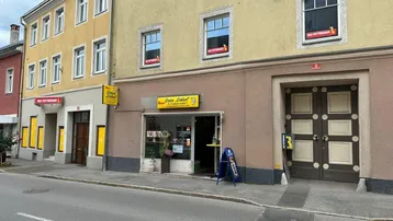 Fassade eines Mehrfamilienhauses mit Ladenlokalen im Erdgeschoss und Wohnungen darüber, an einer Straße gelegen.