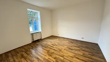 Helles, leeres Zimmer mit Holzparkettboden und einem Fenster, das viel Tageslicht hereinlässt.
