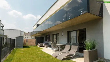 Sonniger Gartenbereich mit Rasenfläche, Liegestühlen und einer Terrasse vor dem modernen Gebäude.