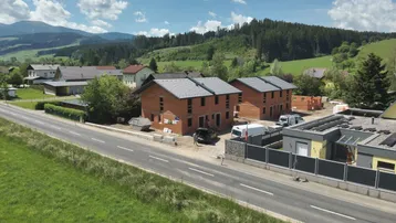 Luftaufnahme einer ländlichen Wohngegend mit Neubauten und grünen Hügeln unter blauem Himmel.