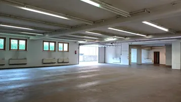 Großer, leerer Lagerraum mit Betonboden, vielen Fenstern und Leuchtstoffröhren an der Decke.