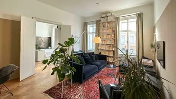 Helles Wohnzimmer mit Fischgrätparkett, hohen Decken und Blick auf die Straße, verbunden mit einem weiteren Raum.