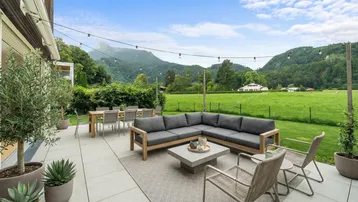 Geräumige Terrasse mit Lounge-Möbeln und Esstisch, umgeben von Bergen und grünen Wiesen.