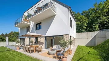 Moderne Hausfassade mit großer möblierter Terrasse, Garten und Balkonen im Obergeschoss.