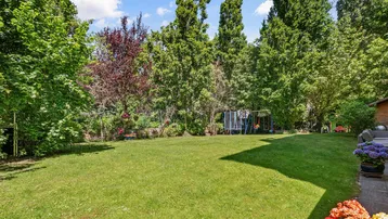 Großer, gepflegter Garten mit weitläufiger Rasenfläche, hohen Bäumen und Spielgeräten für Kinder.