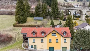 Traditionelles Haus mit rotem Ziegeldach und grüner Umgebung, umgeben von Bäumen und Bergen.