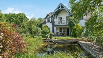Großzügiges Anwesen mit Teich, Holzsteg und üppiger Gartenanlage unter blauem Himmel.