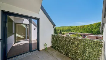 Heller Balkon mit Fliesenboden und weitem Blick ins Grüne und auf bewaldete Hügel.