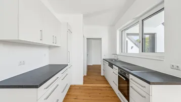 Moderne Küche mit weißen Schränken, dunklen Arbeitsplatten und Holzboden, gut beleuchtet durch ein Fenster.