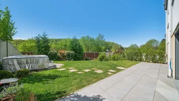 Großer Garten mit Rasenfläche, Whirlpool und gepflasterter Terrasse, umgeben von viel Grün.