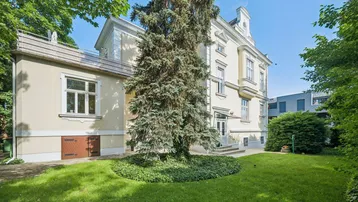 Elegantes Altbaugebäude mit gepflegtem Vorgarten und altem Baumbestand unter blauem Himmel.