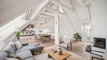 Helles Dachgeschoss-Wohnzimmer mit sichtbaren Holzbalken, Kamin und offenem Essbereich.