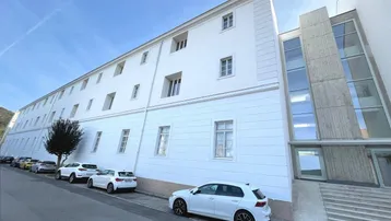 Weißes Gebäude mit moderner Glasfassade und parkenden Autos auf der Straße.