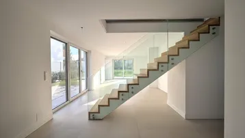 Geräumiger Wohnbereich mit großen Fenstern, Fliesenboden und moderner Holz-Glas-Treppe.