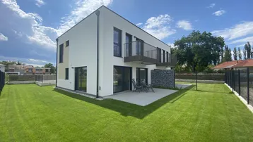 Modernes Einfamilienhaus mit weißer Fassade, großem Rasen und möblierter Terrasse unter blauem Himmel.