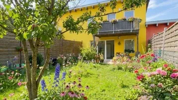 Gepflegter Garten mit blühenden Blumen und Rasenfläche vor einem gelben Haus mit Balkon.