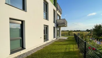 Moderne Gebäudefassade mit Balkonen, Rasenfläche und Blick ins Grüne unter blauem Himmel.