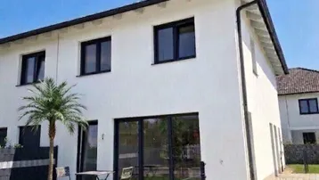 Modernes Einfamilienhaus mit weißer Fassade, großen Fenstern, kleiner Terrasse und gepflegtem Gartenbereich.