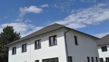Modernes Einfamilienhaus mit weißer Fassade, dunklem Dach und gepflasterter Einfahrt unter blauem Himmel.
