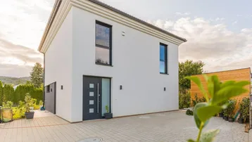 Modernes Einfamilienhaus mit weißer Fassade, dunklen Fenstern und gepflasterter Auffahrt.