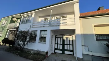 Weißes Gebäude mit grünem Garagentor und einem Balkon im Obergeschoss, umgeben von einer kleinen Gartenfläche.