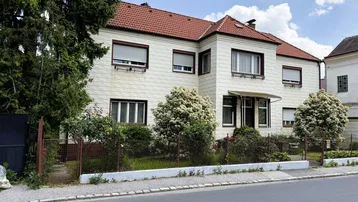 Einfamilienhaus mit rotem Ziegeldach, heller Fassade und Vorgarten an einer ruhigen Straße.