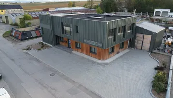 Modernes Gebäude mit dunkler Fassade und Holzverkleidung, umgeben von gepflasterten Flächen und ländlicher Umgebung.