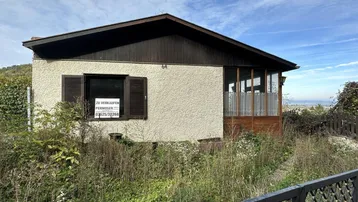 Einfaches Einfamilienhaus mit Putzfassade und Holzverkleidung, umgeben von üppiger Vegetation.