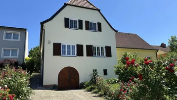 Traditionelles Haus mit heller Fassade, braunen Fensterläden und einem Vorgarten voller Rosen.