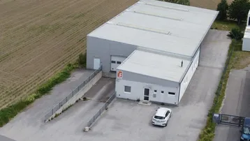 Luftaufnahme eines modernen Gewerbegebäudes mit Lagerhalle und Bürotrakt, umgeben von Feldern und Parkplätzen.