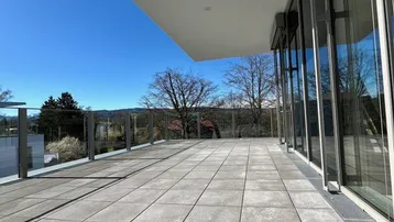 Große, sonnige Terrasse mit modernen Fliesen, Glasgeländer und weitem Blick ins Grüne.