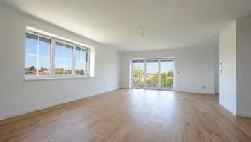 Geräumiges Wohnzimmer mit hellem Holzboden, großen Fenstern und Zugang zu einem Außenbereich.