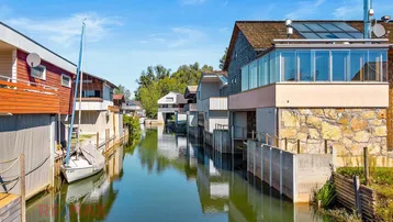 Moderne Häuser am Kanal mit Bootsanlegestellen und einem Segelboot, umgeben von grüner Natur.