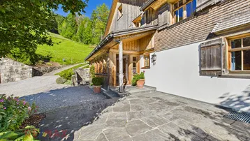 Traditionelles Holzhaus mit Steinfliesen am Eingangsbereich und grüner Hanglage.