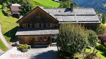 Traditionelles Holzhaus mit Schindeldach, umgeben von grünen Hügeln und Bäumen in alpiner Landschaft.