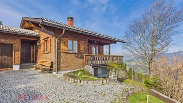 Traditionelles Holzhaus mit Balkon und gepflasterter Auffahrt in alpiner Umgebung.