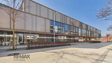 Moderne Geschäftsfassade mit großen Glasflächen und umliegendem Gehweg, umgeben von Bäumen unter blauem Himmel.