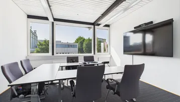 Moderner Konferenzraum mit großem Tisch, bequemen Stühlen und einem Wandfernseher für Präsentationen. Große Fenster bieten viel Tageslicht.