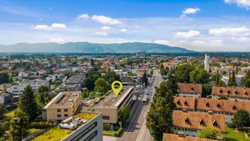 Luftaufnahme einer Stadt mit Bergen im Hintergrund und einem markierten Gebäude unter blauem Himmel.