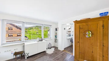 Heller Wohnbereich mit Fensterfront, Kommode und Blick auf die umliegenden Häuser und Hügel.