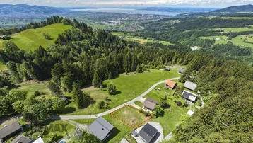 Luftaufnahme eines Baugrundstücks in hügeliger Landschaft mit Blick auf den See und umliegende Häuser.