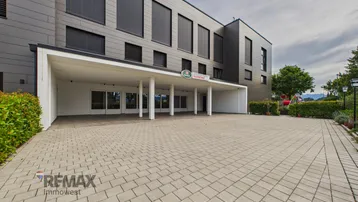 Moderne Gebäudefassade mit überdachtem Eingangsbereich und großem gepflasterten Vorplatz.