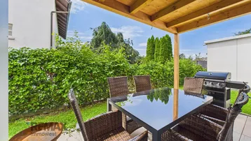 Überdachte Terrasse mit Esstisch, Stühlen und Grill, umgeben von einer grünen Hecke.