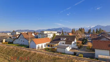 Luftaufnahme eines Wohnhauses mit angrenzenden Bahngleisen und Bergpanorama im Hintergrund.